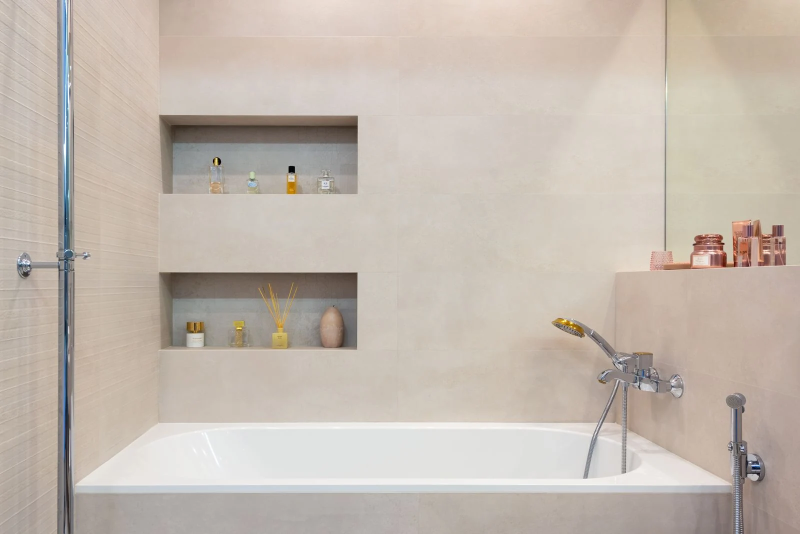 Interior of light modern bathroom with white bath with shower under shelves with decorative objects next to mirror